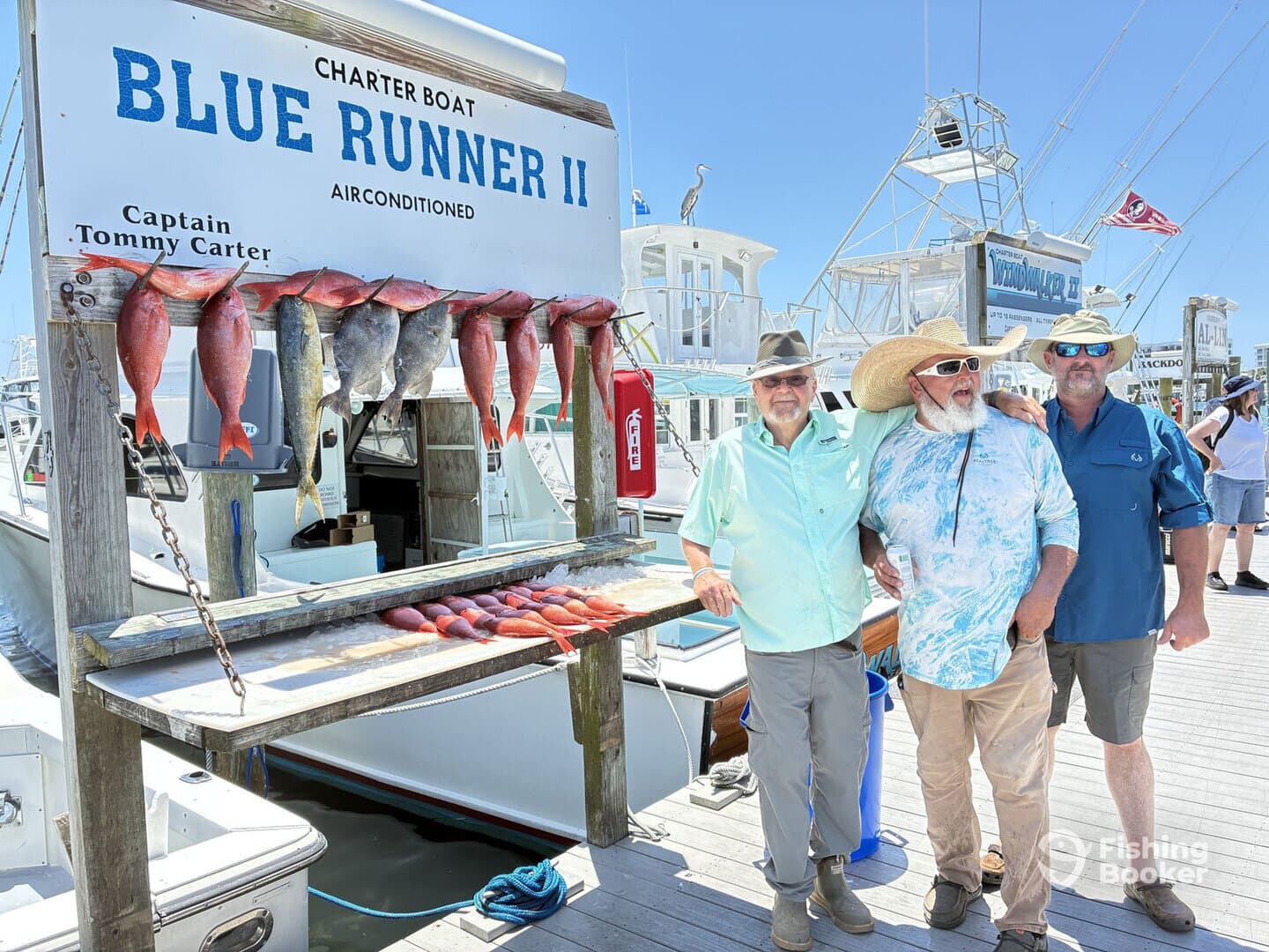 Three anglers posing in front of a catch board displaying a variety of fish, including snapper and mahi-mahi, at a charter boat dock.