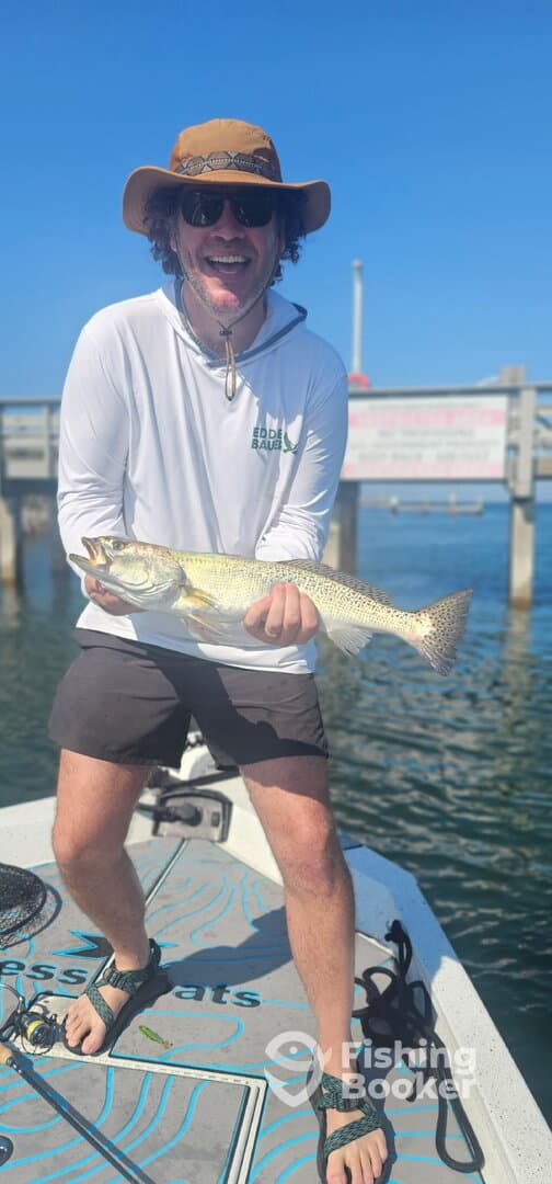 Angler proudly displaying a large Speckled Trout while standing on a fishing boat, enjoying a sunny day on the water.
