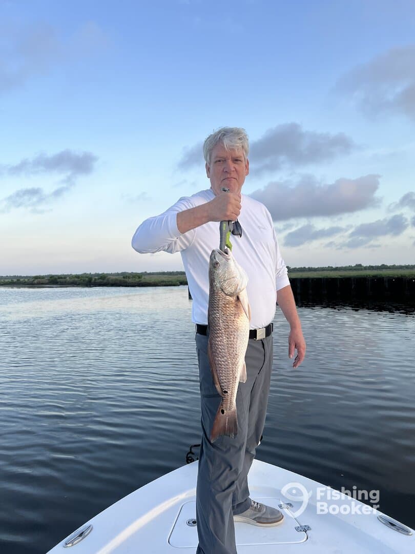 Angler proudly displaying a large Redfish while standing on the bow of a boat during a serene fishing trip.