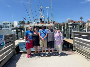 A group of five anglers posing in front of their charter boat at the marina, showcasing a sunny day and a vibrant fishing atmosphere.