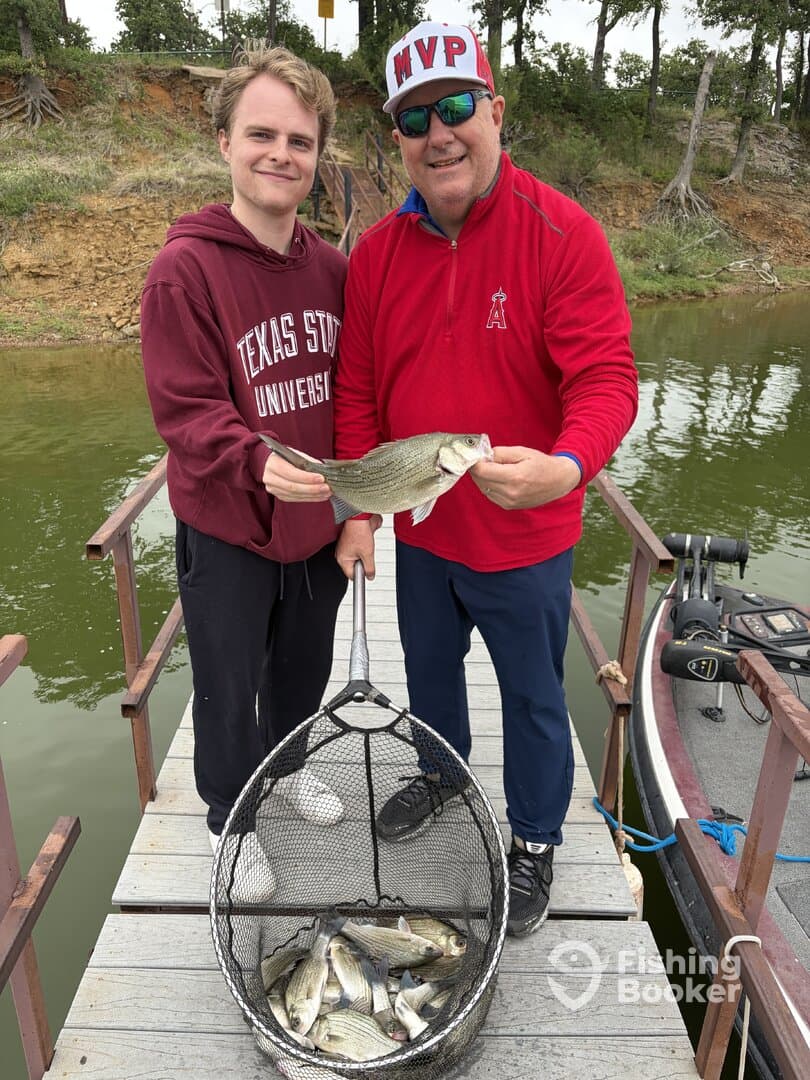 Two anglers proudly displaying a large Striped Bass caught during a fishing trip, with a net full of additional fish visible.