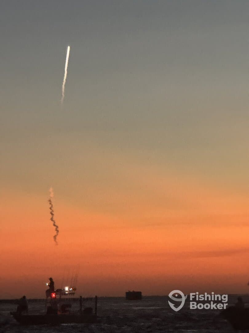 A fishing boat silhouetted against a colorful sunset, with a rocket trail visible in the sky.