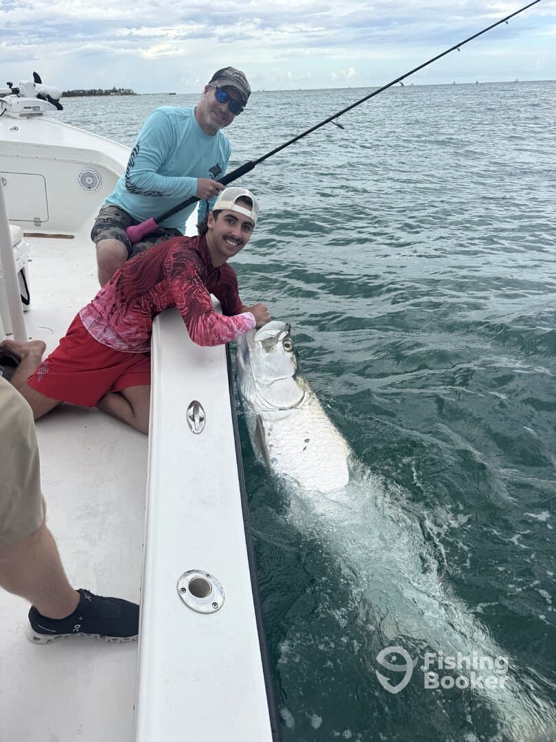 Two anglers engaged in the excitement of reeling in a large Tarpon while fishing from a boat in coastal waters.