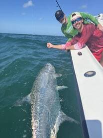 Two anglers actively reeling in a large Tarpon while fishing in open waters, showcasing the excitement of sport fishing.