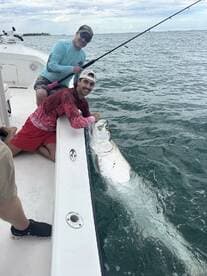 Two anglers engaged in the excitement of reeling in a large Tarpon while fishing from a boat in coastal waters.