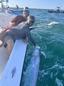 Two anglers actively reeling in a large Tarpon while fishing in clear blue waters, showcasing an exciting moment on the boat.