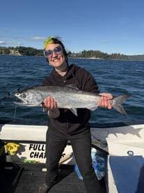 Angler proudly displaying a large Salmon caught during a fishing trip, with a scenic backdrop of trees and water.