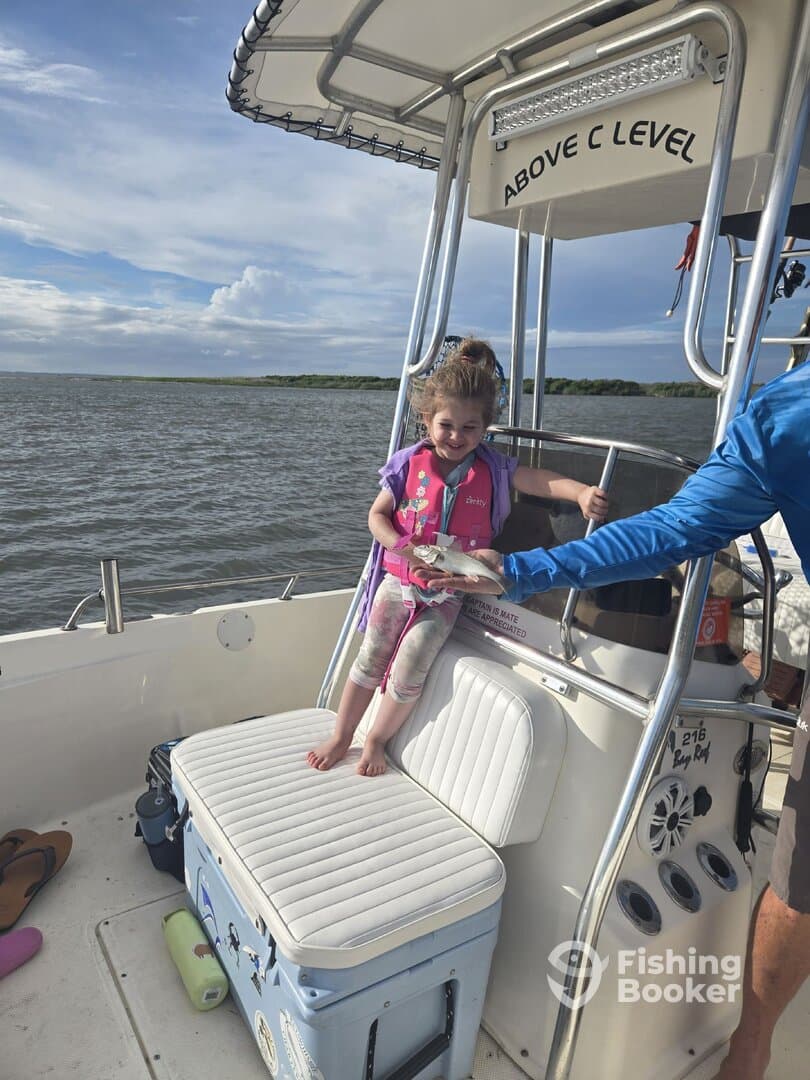 A young girl in a life jacket joyfully holds a small fish while sitting on a cooler aboard a fishing boat, enjoying a day on the water.