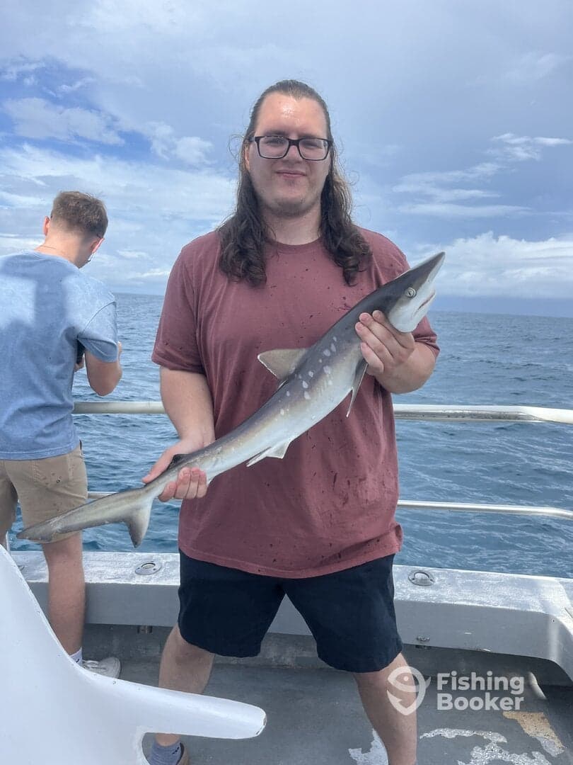 Angler proudly displaying a caught shark while fishing on a boat, with another person in the background.