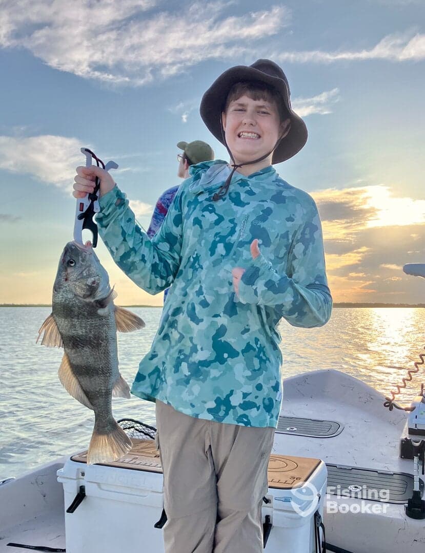 A young angler proudly displaying a large Black Bass while fishing on a boat during sunset.