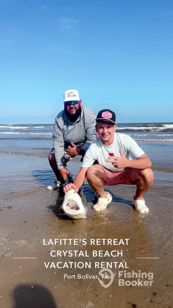 Two anglers proudly displaying a caught shark on the beach at Port Bolivar, TX, showcasing a fun fishing experience.