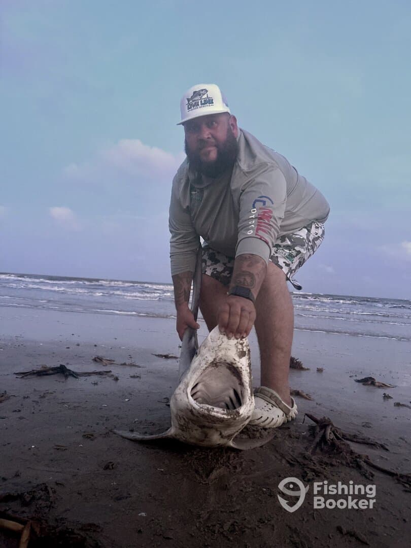 Angler proudly displaying a large shark on the beach, showcasing a successful catch during a coastal fishing trip.
