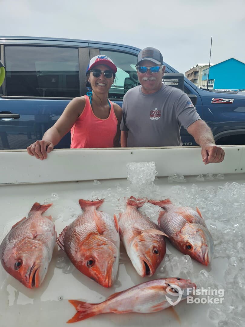 Two anglers proudly displaying their catch of five Red Snapper on a fishing trip, with a vibrant backdrop of a dock and vehicles.