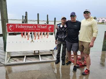 Three anglers posing in front of a catch board displaying a variety of fish, including Red Snapper, at Fisherman's Wharf in Port Aransas, Texas.