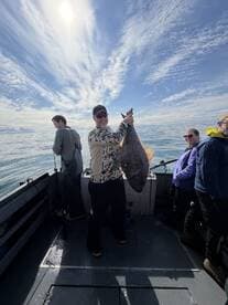 A proud angler displaying a large Halibut while fishing on a boat, surrounded by fellow anglers under a bright sky.