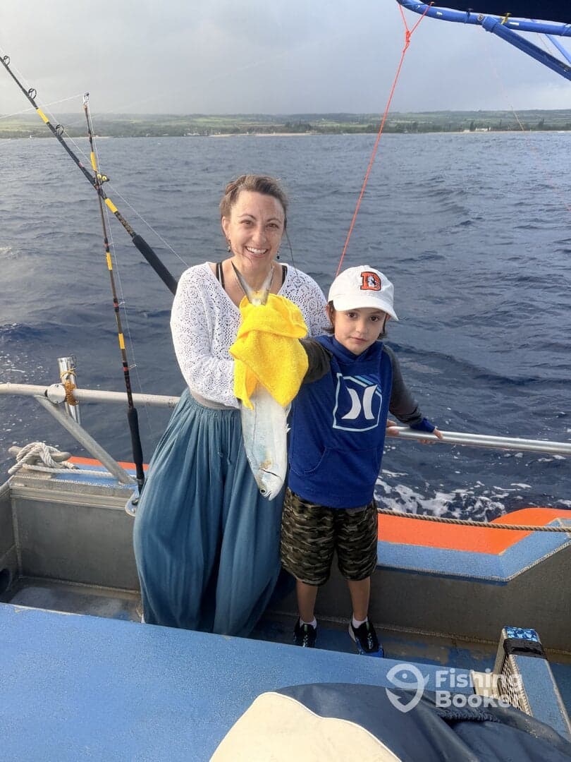 A mother and her young son proudly display their catch on a fishing trip, showcasing a fresh fish against the backdrop of the ocean.