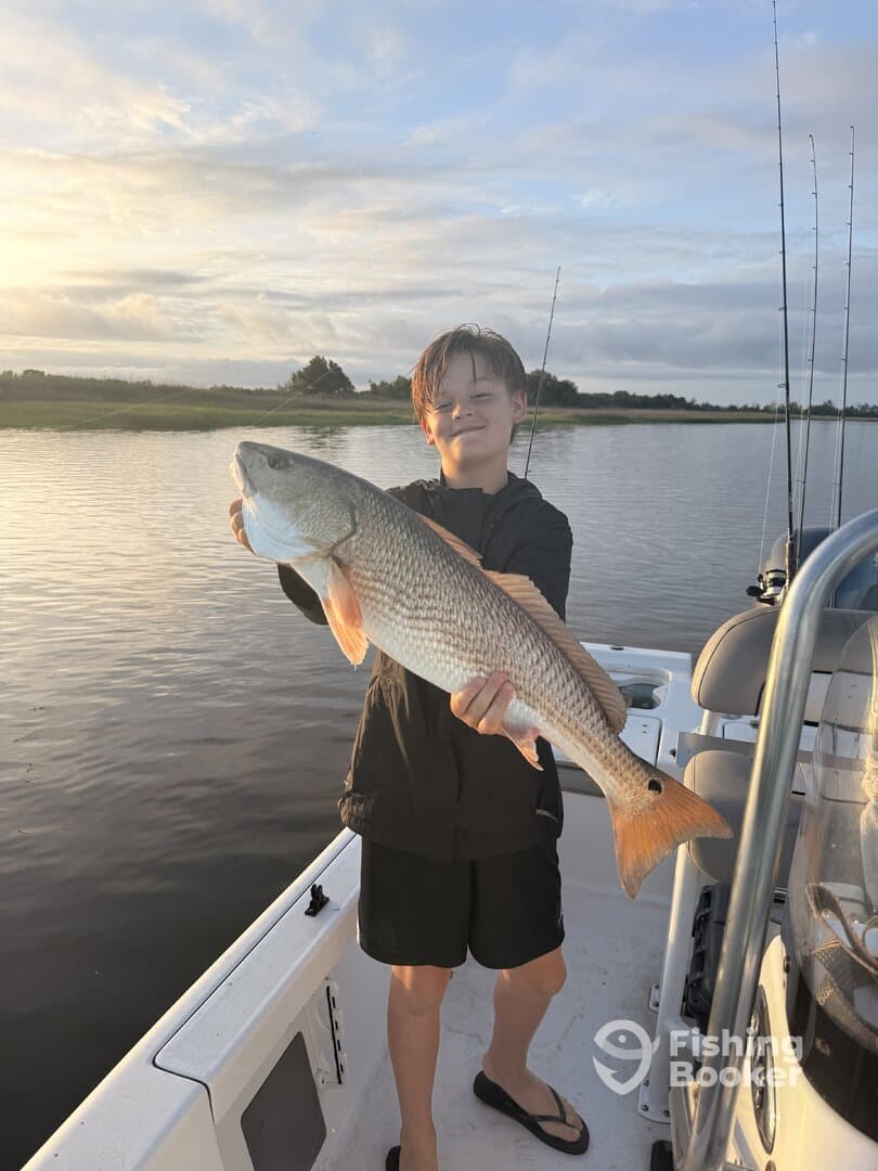 A young angler proudly displaying a large Redfish while fishing on a calm waterway during sunset.
