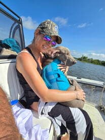 A woman enjoying a relaxing moment on a boat, holding a dog in a life jacket, with a scenic water backdrop.