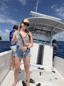 A young woman proudly displaying her catch, a large fish, while aboard a fishing boat in sunny waters.