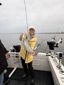 A smiling angler proudly displays a large Brown Trout while fishing on a boat, showcasing a successful catch in a scenic waterway.