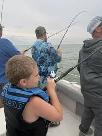 A young angler in a blue life vest is actively reeling in a fish while fishing with a group on a boat in a cloudy environment.