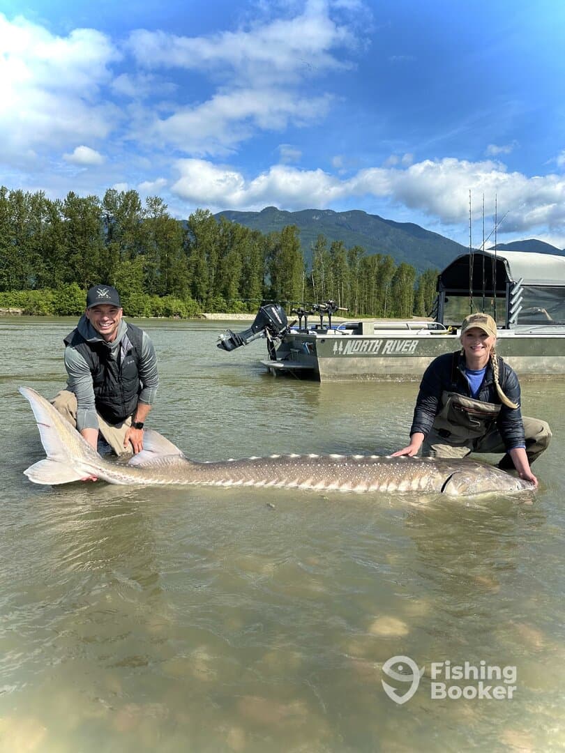 Two anglers proudly displaying a large Sturgeon while standing in shallow water, with a North River boat visible in the background.