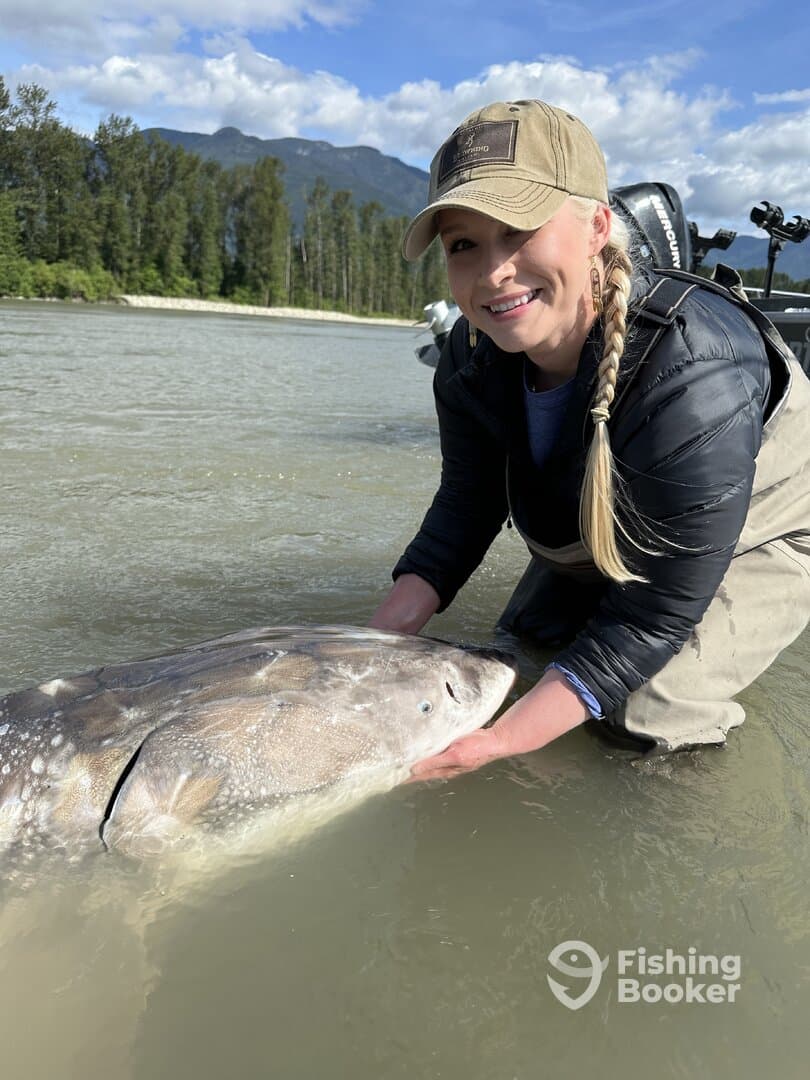 Angler smiling while holding a large Sturgeon in a scenic river setting, showcasing a successful fishing experience.