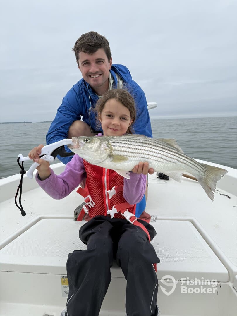 A young girl proudly displays a striped bass while sitting on a boat with an adult, showcasing a memorable fishing experience.