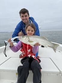 A young girl proudly displays a striped bass while sitting on a boat with an adult, showcasing a memorable fishing experience.