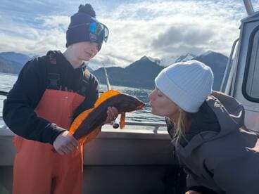 A young angler proudly holding a vibrant rockfish while sharing a playful moment with a companion aboard a fishing boat in a scenic coastal setting.