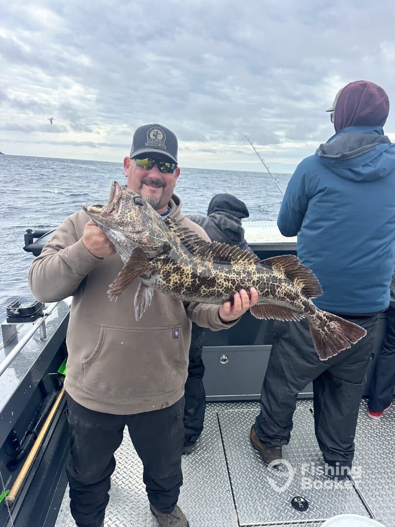 Angler proudly displaying a large Lingcod while fishing on a charter boat, with fellow anglers in the background.