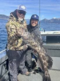 Two anglers proudly displaying a large Lingcod while fishing in a scenic coastal environment.