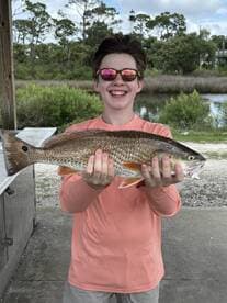 A young angler proudly displaying a Redfish at a scenic dock, showcasing a successful fishing trip.