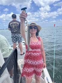 A woman proudly displaying a Speckled Trout while fishing on a boat, enjoying a sunny day on the water.