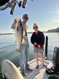Two anglers proudly displaying a large striped bass while fishing on a calm lake during sunrise.
