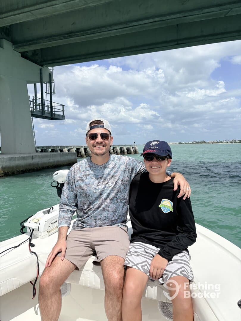 A father and son enjoying a sunny day on a boat, with a scenic view of the water and a bridge in the background.