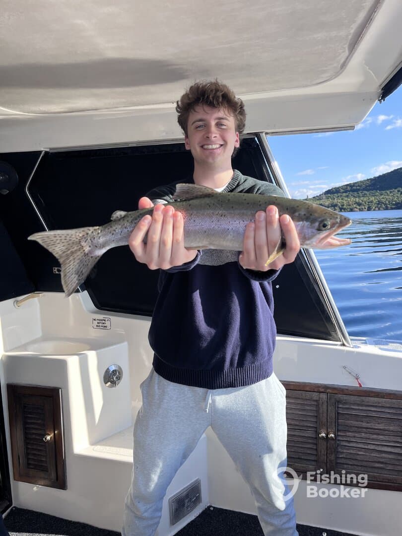 A young angler proudly displaying a Rainbow Trout aboard a fishing boat, showcasing a successful day on the water.