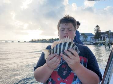 A young angler proudly displaying a striped fish while fishing from a boat, with a scenic waterfront in the background.