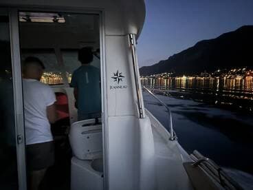 Two individuals inside a Jeanneau boat, navigating through a scenic harbor illuminated by city lights at dusk.