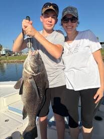 A young angler proudly displaying a large Black Drum while fishing with a family member on a sunny day.