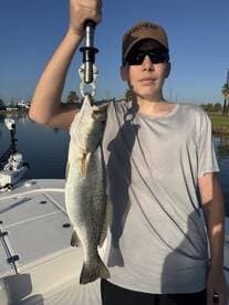 Young angler proudly holding a large Speckled Trout on a boat in a calm fishing environment.