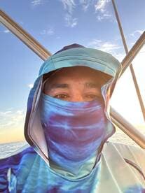 A person wearing a colorful fishing shirt and mask, posing on a boat during sunset.