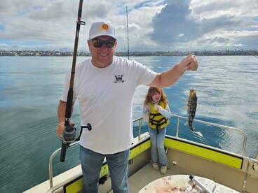 A proud angler holding a freshly caught fish while a young girl in a life jacket joyfully watches on a boat in calm waters.