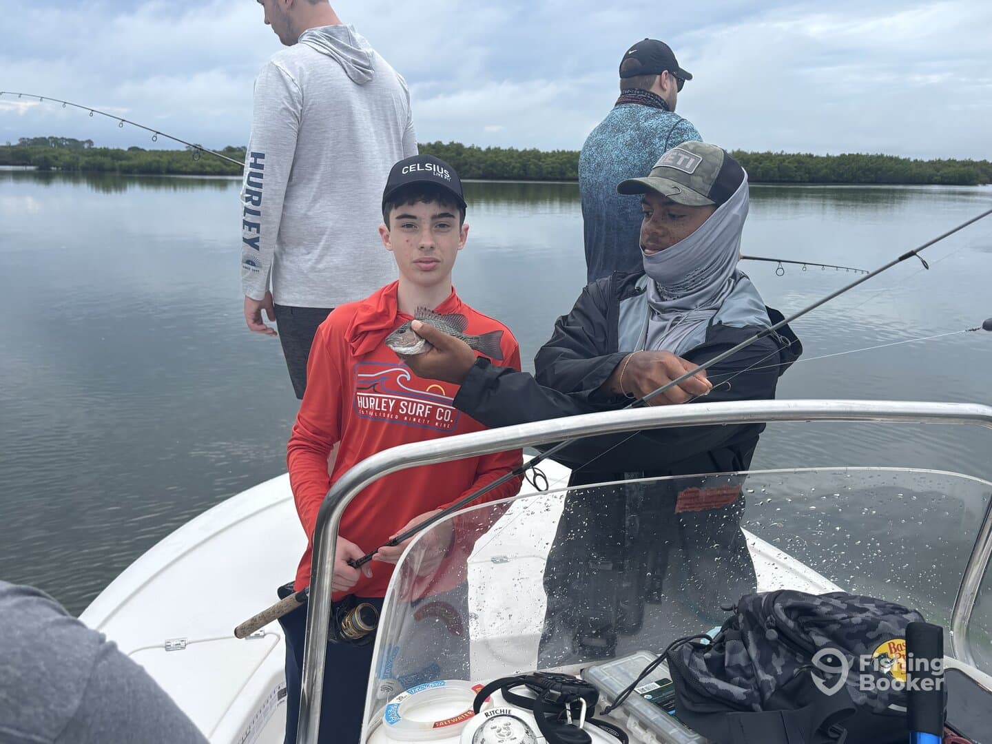 A young angler holds a small fish while being guided by an instructor on a boat, with a scenic backdrop of calm waters and mangroves.