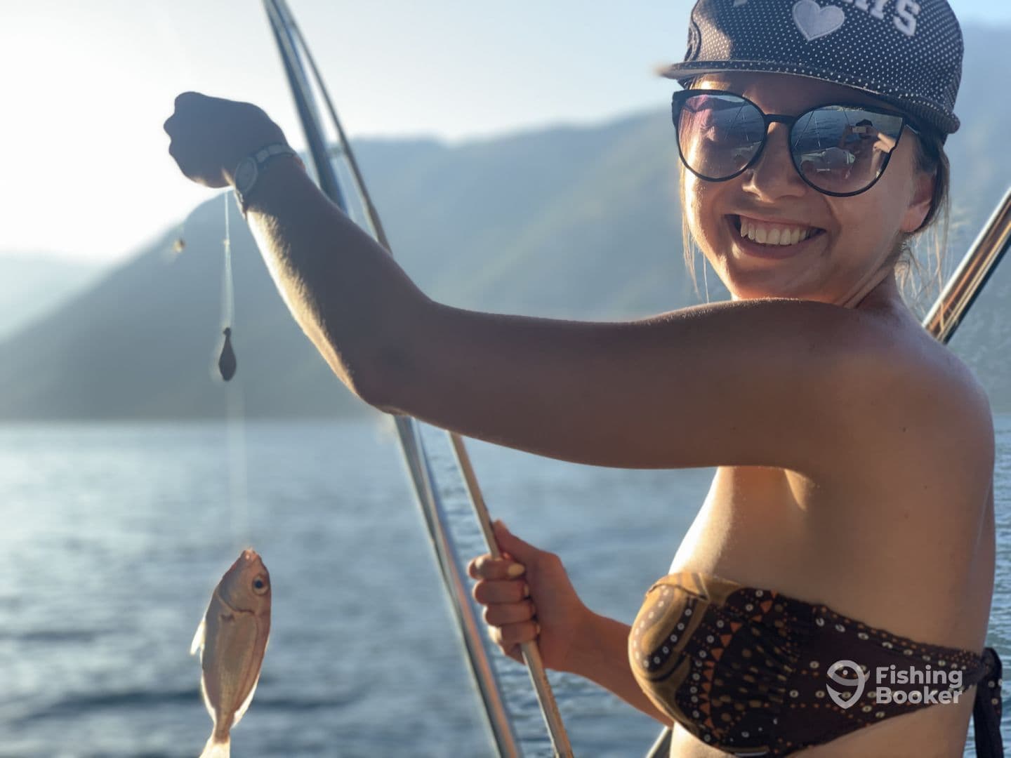 A smiling angler holding a freshly caught fish while fishing on a boat, surrounded by scenic mountains and water.