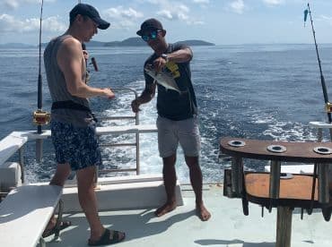 Two anglers on a boat engaged in fishing, with one holding a freshly caught fish while the other prepares to assist. The backdrop features a clear ocean and distant islands.