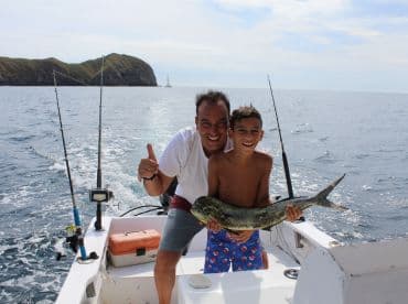 A young boy and his father proudly display a Mahi Mahi while fishing on a boat, surrounded by a scenic ocean backdrop.