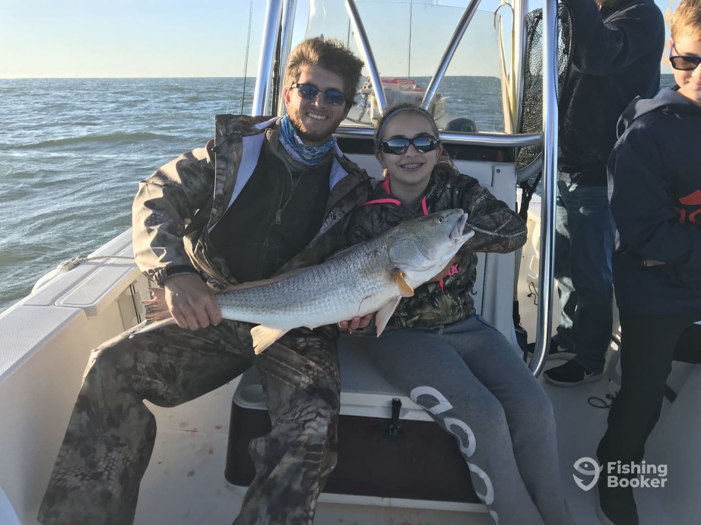 A young angler and a friend proudly displaying a large Redfish aboard a fishing boat, enjoying a day on the water.