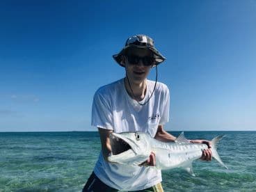 An angler proudly displaying a large Barracuda while standing in shallow waters, showcasing a sunny day fishing experience.