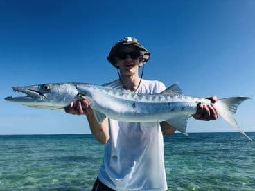 A young angler proudly displaying a large Barracuda while standing in shallow waters, showcasing a successful fishing trip.